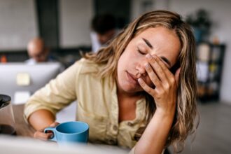 Kimerült nő az irodában, kávéval a kezében, fáradtság jelei az arcán