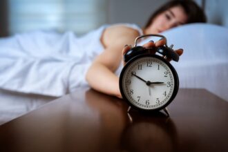 Nő ébren fekszik az ágyban, az előtérben ébresztőóra hajnali 3 órai idővel