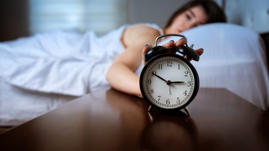 Nő ébren fekszik az ágyban, az előtérben ébresztőóra hajnali 3 órai idővel